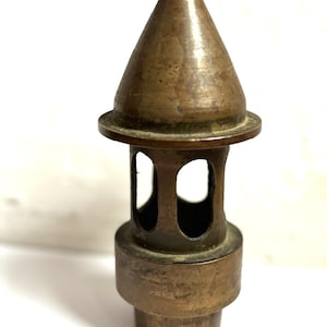 May include: A weathered brass object featuring a conical top section, a central cylindrical body with four arched cutouts, and a solid base. The metal surface displays a dark, aged patina with visible signs of oxidation and wear throughout the piece.