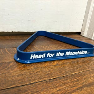May include: A blue pool rack with the text "Head for the Mountains..." in white. The triangular rack is made of metal and sits on a wooden surface. The rack is used to arrange billiard balls.