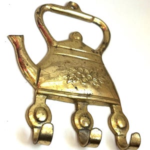 May include: A brass wall hook shaped like a teapot with a handle at the top and three individual hooks extending from the base. The teapot body features an embossed floral design in the center and shows signs of aged patina and surface wear.
