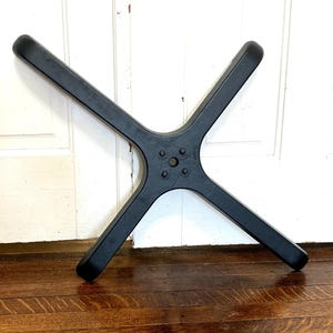 May include: A black, four-legged base, likely for a chair or table, is displayed against a white door. The base has a central hub with four screw holes and rounded ends on each leg. The legs extend outward in a cross shape.