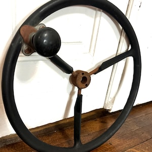 May include: A black steering wheel featuring a three-spoke design with a central metal hub showing signs of rust. A black spherical knob is attached to the upper left section of the rim, secured by a metal bracket with visible oxidation.