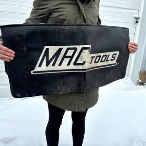 May include: A black tool bag with the white text "MAC TOOLS" is held in front of a person. The bag is rectangular and appears to be made of a durable material. The background includes a white garage door and snow.