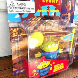 Może przedstawiać: Figurka akcji Alien z Toy Story w przezroczystym plastikowym opakowaniu. Zielony kosmita ma trzy oczy, niebieski mundur i ż&oacute;łtą głowę. Opakowanie zawiera napis "Toy Story" i ostrzeżenie o niebezpieczeństwie uduszenia.