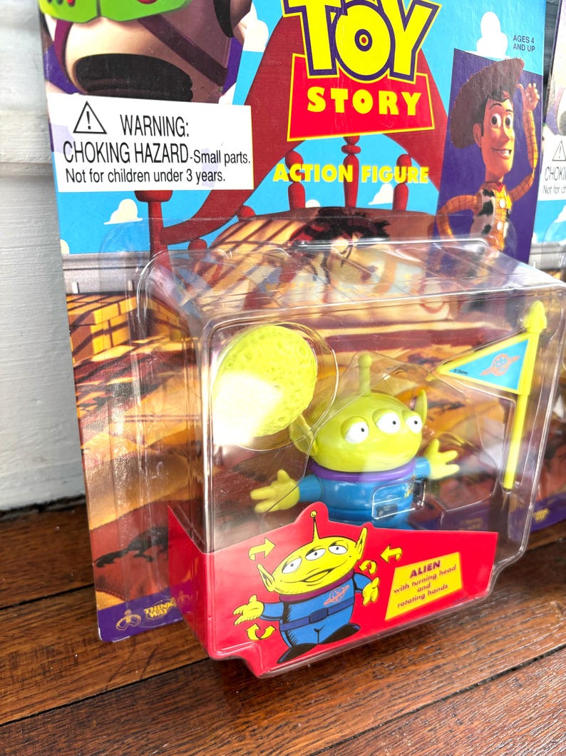 Może przedstawiać: Figurka akcji Alien z Toy Story w przezroczystym plastikowym opakowaniu. Zielony kosmita ma trzy oczy, niebieski mundur i ż&oacute;łtą głowę. Opakowanie zawiera napis "Toy Story" i ostrzeżenie o niebezpieczeństwie uduszenia.
