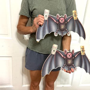 May include: Two identical die-cut cardboard signs featuring a cartoon bat with dark purple wings and a furry body. The bat is depicted holding a Coors Light beer can in each clawed hand. The signs have a glossy finish and a hanging hole at the top.