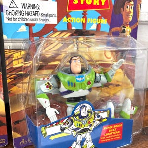 Może przedstawiać: Figurka Buzz Lightyear w oryginalnym opakowaniu. Figurka jest zielona, biała i fioletowa, z przezroczystym plastikowym bąblem. Opakowanie zawiera logo Toy Story i ostrzeżenie o małych częściach.
