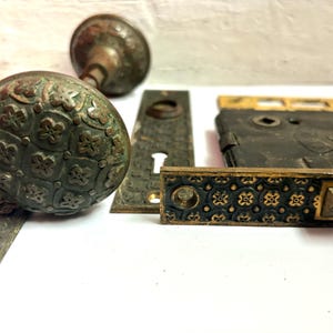 May include: Antique bronze door hardware set, including a doorknob with a floral pattern, a matching backplate, and a mortise lock. The hardware has a weathered appearance, suggesting age and use. The set is likely from the early 20th century.