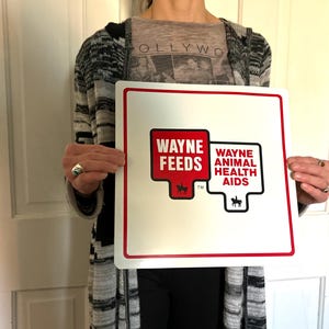 May include: Square white sign with red and white text that reads "WAYNE FEEDS" and "WAYNE ANIMAL HEALTH AIDS". The sign has a red border and a black silhouette of a horse. The sign is held by a person.