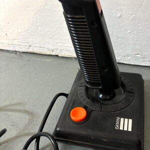 May include: A black video game joystick with a vertical handle, a bright orange top button, and a matching orange base button. The base features the word GEMINI printed in white text next to three white vertical bars and a black coiled cord.