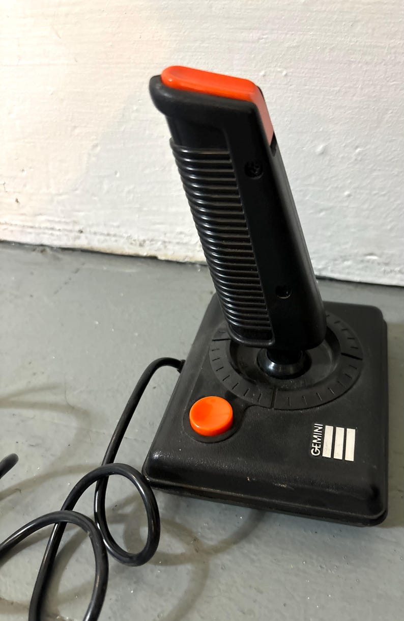May include: A black video game joystick with a vertical handle, a bright orange top button, and a matching orange base button. The base features the word GEMINI printed in white text next to three white vertical bars and a black coiled cord.
