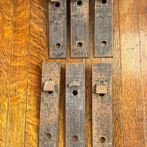 May include: Six rectangular metal door plates with a dark, weathered finish. Each plate features an intricate embossed pattern including floral motifs, geometric borders, and a central shield design, with a small lever and two circular openings.