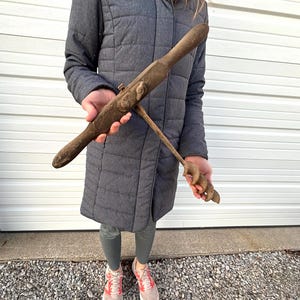 May include: Antique wooden and metal tool with a long, tapered handle and a curved, spiral-shaped end. The tool is held against a gray coat and appears to be weathered, suggesting age and use. The tool's design indicates it may have been used for woodworking or similar crafts.