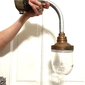 May include: A wall-mounted industrial light fixture featuring a curved silver metal conduit arm, a weathered dark gray junction box, and a copper-toned metal socket housing. A clear, rounded glass globe shade is attached to the copper socket base.