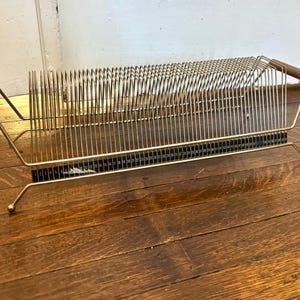 May include: A gold-toned metal record rack featuring a series of vertical wire dividers for organizing vinyl albums. The rack includes two cylindrical wooden handles on each end and a black base strip with white numbered markings from one to fifty.