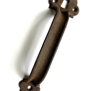 May include: A rustic, brown metal handle with a rectangular grip and decorative ends. Each end features a heart-shaped design with a circular hole and a square opening in the center. The handle is likely for a cabinet or drawer.