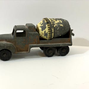 May include: A vintage toy cement mixer truck made of weathered gray metal with visible rust patches. The truck features a rotating drum painted in a distressed yellow and gray finish, six black wheels, and an open cab design with no windows.