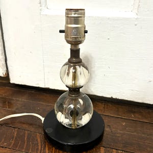 May include: A vintage table lamp with a black base and a clear, spherical glass body. The lamp features two stacked glass spheres and a metal lamp holder. A white electrical cord extends from the base.