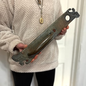 May include: A long, rectangular, weathered brass door plate featuring an integrated handle and ornate, scrolled leaf-like detailing at both the top and bottom ends. The metal surface shows a dark, aged patina with a circular hole at the top.