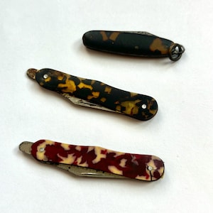 May include: Three small pocket knives with decorative handles. One has a black and brown tortoiseshell pattern, another has a red and cream pattern, and the third has a dark green and brown pattern. Each knife has a silver-colored blade.