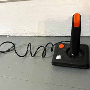 May include: A black video game joystick with a rectangular base, a vertical handle, and an orange fire button on the top and side. The base features the white text GEMINI and three horizontal lines. A black cord with a connector is attached.
