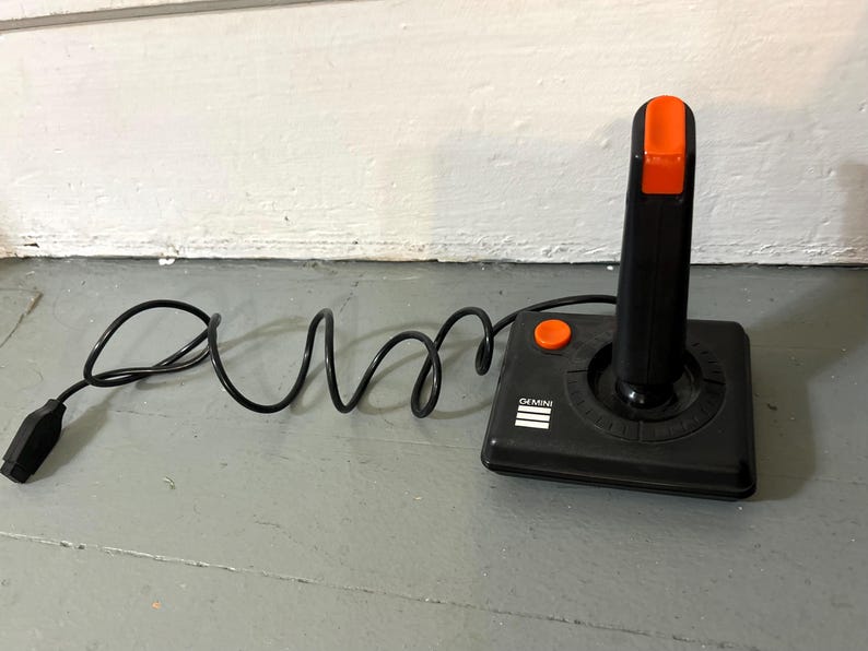 May include: A black video game joystick with a rectangular base, a vertical handle, and an orange fire button on the top and side. The base features the white text GEMINI and three horizontal lines. A black cord with a connector is attached.