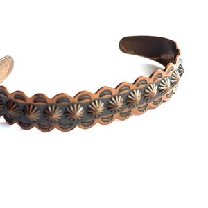 May include: A copper cuff bracelet with a scalloped edge and a repeating pattern of starburst designs. The open-ended bracelet has a rustic, aged appearance, with a dark patina in the recessed areas of the design.