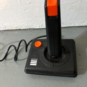 May include: A black video game joystick with a bright orange fire button on the top and a smaller orange button on the base. The base features the white text GEMINI above three horizontal white lines. A black cord extends from the side of the unit.