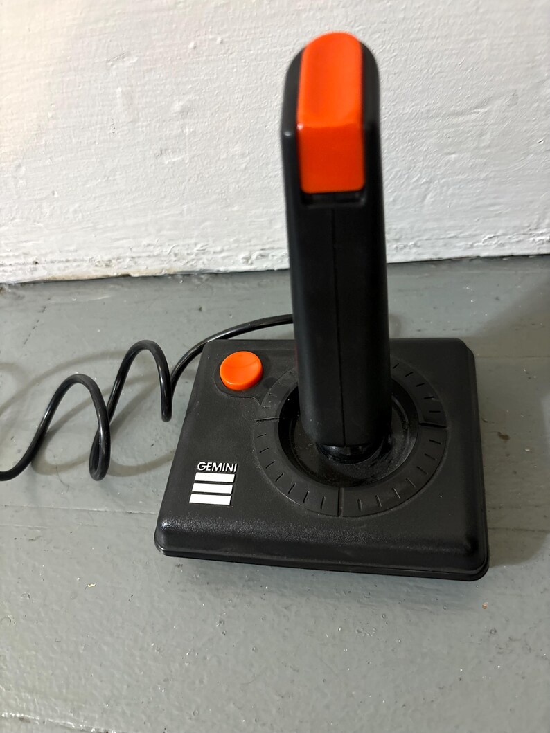May include: A black video game joystick with a bright orange fire button on the top and a smaller orange button on the base. The base features the white text GEMINI above three horizontal white lines. A black cord extends from the side of the unit.