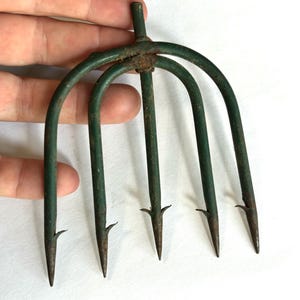 May include: A five-pronged metal fishing spear or gig with a weathered green painted finish and visible rust spots. The tool features five sharp, pointed tines, each equipped with a small barb near the tip, and a central cylindrical attachment point.