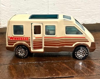 May include: A vintage toy Winnebago RV in cream, brown, and red. The toy RV has black tires with the word "Buddy L" on the sidewalls. The Winnebago logo is visible on the side of the vehicle. The toy has a dark blue sunroof.