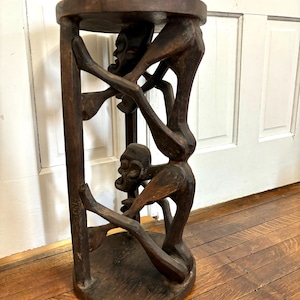 May include: A dark brown wooden stool featuring a circular top and base connected by three carved figures. The figures are depicted in a seated, crouched position with their limbs forming the structural support between the top and bottom.