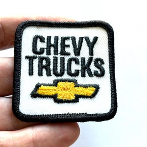 May include: A square embroidered patch with a white background and a thick black border. The text CHEVY TRUCKS is stitched in black capital letters above a yellow Chevrolet bowtie logo, which is outlined in black thread.