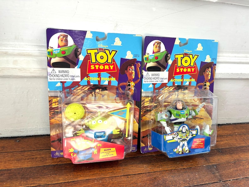 Może przedstawiać: Dwie figurki akcji Toy Story w oryginalnym opakowaniu. Jedna przedstawia Buzza Astrala, druga Obcego. Opakowanie zawiera logo Toy Story, zdjęcia postaci i ostrzeżenie o niebezpieczeństwie uduszenia.