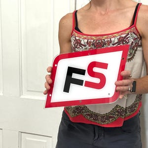 May include: A red and white sign with the letters "FS" in black and red. The sign has a clear, rounded border and is held in front of a white door.