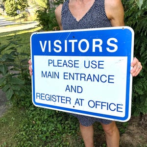 May include: A rectangular blue and white metal sign with rounded corners. The sign features the text VISITORS in white on a blue background, followed by the text PLEASE USE MAIN ENTRANCE AND REGISTER AT OFFICE in blue on a white background.