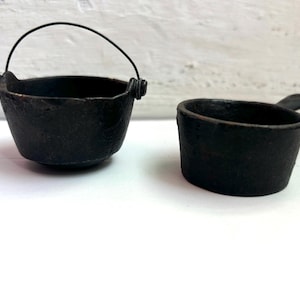 May include: Two small, black cast iron pots. One is a cauldron shape with a handle, and the other is a small cup with a handle. The pots have a textured surface and are set against a white background.