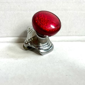 May include: A vintage-style steering wheel spinner knob featuring a circular, translucent red top with a textured, reflective surface. The base is constructed from polished chrome-colored metal with a tiered, stepped design and a mounting clamp.