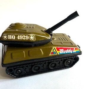 May include: Olive green toy tank with a black cannon and black treads. The tank has the text "HQ 4929" and a "Buddy L" logo. A classic toy vehicle.