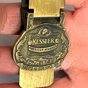 May include: Vintage brass money clip with the Kessler American Whiskey logo. The clip features the brand name, a skull, and the phrase "Smooth as Silk Since 1888". The clip is held in a hand.