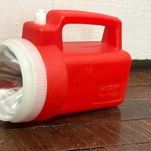 May include: A vintage red flashlight with a white bezel and handle. The flashlight has a clear lens and a white on/off switch. The body of the flashlight is red plastic. The flashlight is made in the USA.