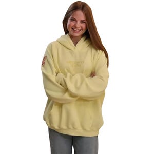 May include: A pale yellow hoodie with the text "SINCERELY YOUNG" embroidered on the front. The hoodie has a hood and long sleeves. A small patch is visible on the left sleeve. The person in the image is wearing blue jeans.