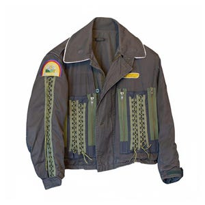 Op de afbeelding: Een donkergrijze jas met een wit afgezette kraag, een regenboogpatch en meerdere ritsen. De jas heeft olijfgroene accenten, waaronder decoratieve veters en ritssluitingsdetails. De jas heeft een knoopsluiting en een gele patch.