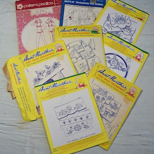May include: A collection of yellow, blue, and pink iron-on transfer patterns for crafts. The patterns feature various designs, including baskets, birds, flowers, and a butterfly. The text on the patterns reads "Aunt Martha's Hot Iron Transfers" and "Each Design Good For Several Stampings".