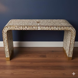 May include: A decorative, curved-front table with a white and brown leaf pattern on the top and sides. The table has a gold-coloured trim and base, and is set against a dark blue wall and brown wood floor.