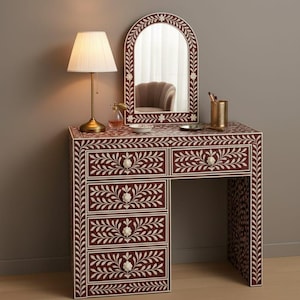 Könnte beinhalten: Burgunderfarbener Schminktisch mit passendem Spiegel, verziert mit einem weißen floralen Intarsienmuster. Der Tisch hat Schubladen und einen Schreibtischbereich. Eine goldene Lampe und Accessoires vervollständigen das elegante Erscheinungsbild.