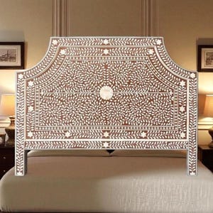 Op de afbeelding: Een decoratief hoofdbord met een bruine achtergrond en witte bloemeninleg. Het hoofdbord heeft een gebogen bovenkant en wordt ondersteund door twee poten. Het hoofdbord is geplaatst boven een bed met een beige sprei. De kamer heeft twee lampen en twee ingelijste foto's.