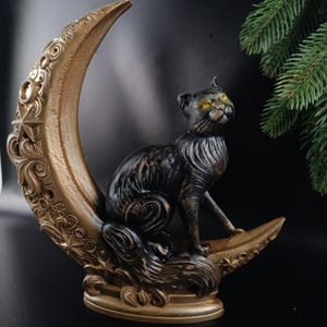 May include: A black cat statue with golden eyes sits on a gold crescent moon. The moon has ornate detailing and stars. The cat is black with bronze highlights. The statue is a decorative item.