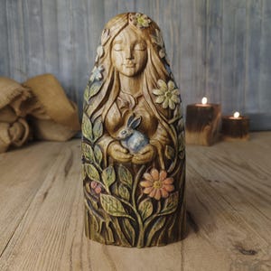 May include: A hand-carved wooden sculpture of a figure with long hair, eyes closed, holding a small rabbit. The figure is surrounded by carved leaves and flowers with subtle green, yellow, and pink paint accents on the natural wood grain surface.