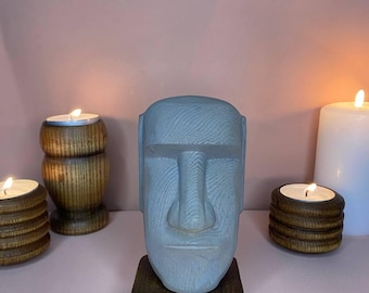 Könnte beinhalten: Eine hellblaue Moai-Statue auf einem dunkelbraunen Holzsockel. Die Statue hat geschnitzte Gesichtszüge und ist von brennenden Kerzen in Holzhaltern umgeben, was eine ruhige Atmosphäre schafft. Der Hintergrund ist ein zartes Rosa.