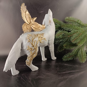 May include: A white wolf figurine with a gold-colored eagle attached to its back. The wolf is howling, and gold-colored tree-like designs adorn its side. A sprig of green pine needles is in the background.